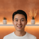 A smiling young adult of East Asian descent looks directly at the camera, a warm amber and terracotta background behind them.